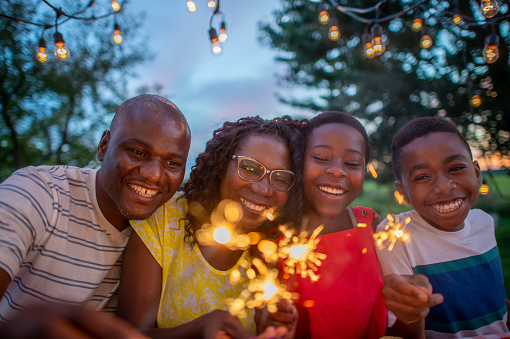 A small African family of four celebrate the 4th of July together in their backyard.  They are each dressed casually and have sparklers in their hands as they smile and enjoy each others company.