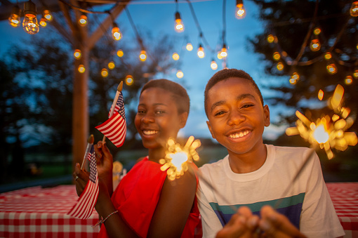 A young brother and sister, of African decent, celebrate the 4th of July together in their backyard.  They are each dressed casually and have sparklers in their hands as they smile and enjoy each others company.