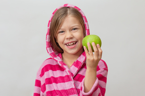 Portrait of caucasian little girl in bathrobe with hood on head holding green apple looking at camera on white background