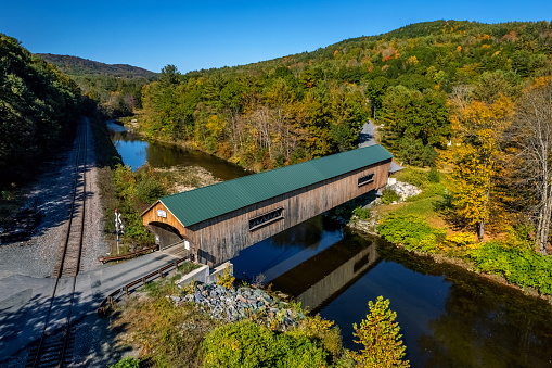 The Bartonsville covered bridge spans a tranquil river, reflecting softly on the water surface, amidst a picturesque, wooded landscape. A parallel railroad track hints at historical means of transport that likely complemented the bridge's role in the area.