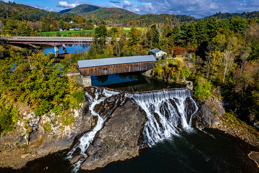 A scenic view showcases a waterfall cascading into a calm pool below, with a covered bridges spanning the river in the background. Lush greenery frames the falls and bridge, set against a backdrop of rolling hills and a clear sky with scattered clouds.