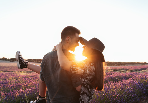 A young couple in love celebrates their anniversary with a romantic date in a lavender field at sunset. The guy picks up his girlfriend in his arms, and they walk among the flowers, looking at each other and smiling.