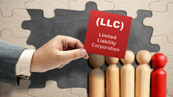 Business hand holding a note with LLC text, the acronym of Limited Liability Company with a row of wooden peg dolls on the table. Business concept
