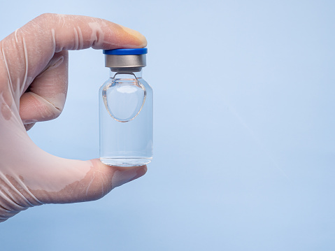 The doctor's hand is in a glove, holding a vaccine vial on a blue background. Close-up. Medical and vaccination concepts.