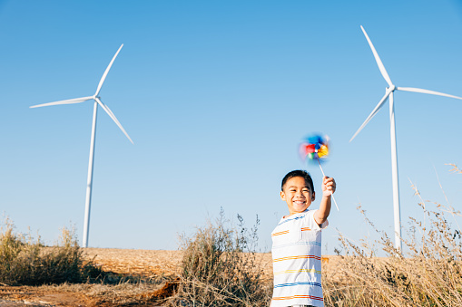 Amidst wind turbines playful boy with pinwheel toy symbolizes excitement of wind energy exploration. Showcases clean electricity innovation in a countryside windmill setting against a serene blue sky.