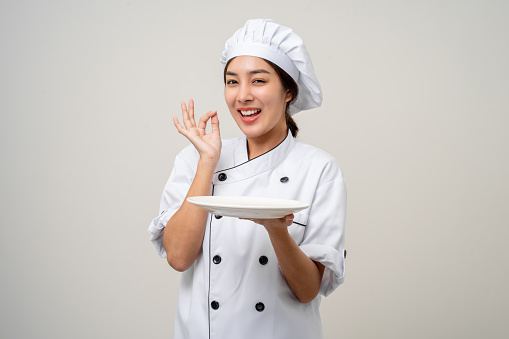 Smiling Young beautiful asian woman chef in uniform holding empty plate delicious dish menu good taste on isolated. Cooking woman Occupation chef or baker People in kitchen restaurant and hotel.