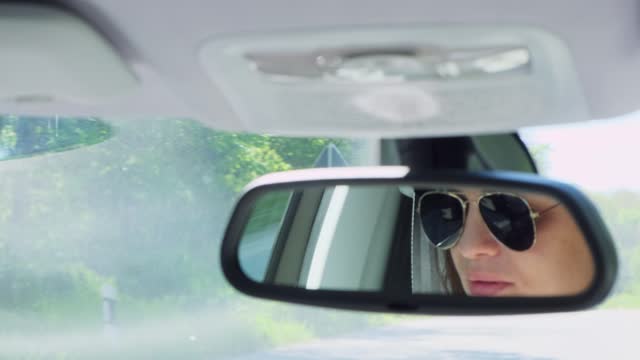 Young confident woman wearing sunglasses driving a car on the highway. Close-up of concentrated female's face reflection in rear view mirror. Road trip concept.
