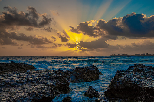 Beautiful sunrise over the sea. Waves crashing onto the rocky floor, with the sun rays and beams of light coming through. Beach and ocean visible in the and the horizon spands into the distance. Suitable for images, printing, wallpaper, desktop background, computer monitor, screensaver.