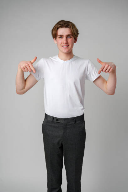 istock Young Man in White T-Shirt Pointing Downwards With Both Hands Against Plain Background 2137326067