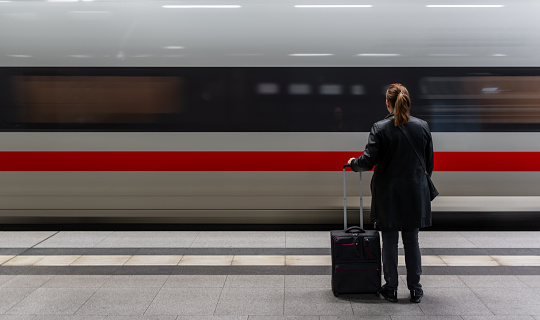 Germany, Berlin, March 26, 2024 -Rear view of woman with luggage standing in front of arriving passenger train, Berlin Hautpbahnhof