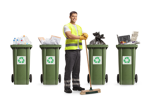 Cleaner in a uniform holding a broom in front of recycling bins isolated on white background