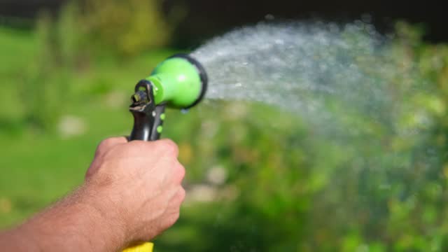 Gardener with a watering hose. Person spraying green grass lawn with hose sprayer. Irrigation with water, sunny day. Garden sprinkler in action. Landscaping. Gardening, waters, growing and plants care