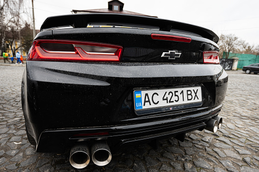 Lutsk, Ukraine - March, 2024: Close-up of a modern black Chevrolet Camaro ZL1 sports car's rear, tail lights, and exhaust pipes parked on a cobblestone street.