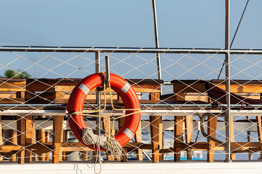 Red lifebuoy with a rope on a metal railing against a blurred ship deck and blue sky background, taken in Side, Manavgat, Turkey