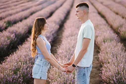 Young couple holding hands together in purple lavender field. Male and female enjoying romantic time in sunset. Beautiful woman hugging man and walking among violet flowers with sunlight on summer day