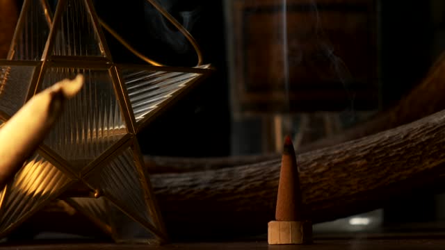 Detail view of a incense cone burning with smoke and a star shaped candle in a dark esoteric setting, in slow motion