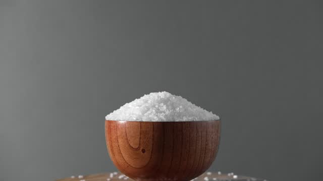White table salt granules falling into a pile. Wood bowl. Green color studio background. Condiment particles dropping. Clean cosmetic bath salt pouring footage. Closeup of grained sea salt pieces hill