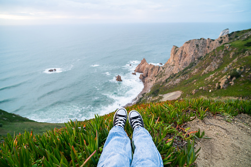 A woman sits high above a serene coastline, legs stretched out. Feet and coastal cliffs. Adventure with tranquility concept. Personal perspective social media style photo. Shot at Cabo da Roca, Portugal.