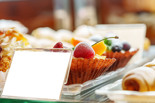 A variety of gourmet desserts, including fruit tarts and pastries, are elegantly presented on a glass display at a catered event, awaiting guests. A blank place card offers the possibility to describe the selection or note allergen information.