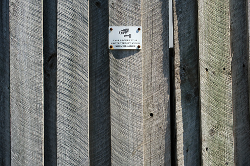 Old metal security camera sign on a wooden fence.
