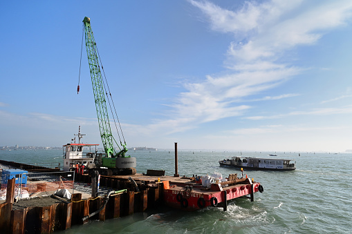 Venice, Italy, march 12, 2024 : Renovation site of a Venice quay with a crane on a barge and a vaporetto in the background