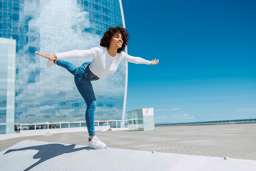 A gorgeous mixed race young smiling woman with curly hair, dancing outdoors at city. Looks like princess maintaining balance with body. Copy space.