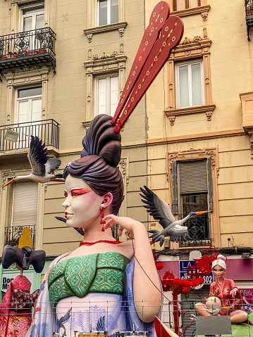 Valencia, Spain - March 19, 2024: Low angle view of a Ninot resembling a geisha in front of old-fashioned building in the city downtown. This are huge dolls that are built specifically to be burnt during the last night of Las Fallas annual event, exclusive to the Valencian Community