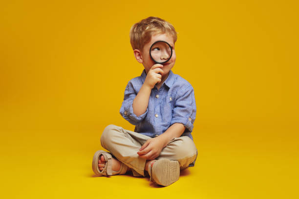 istock Little curious schoolboy sitting on floor with crossed legs while looking away through magnifier. 2137964157