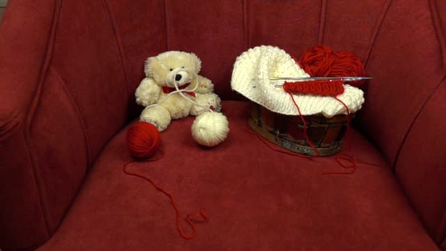Two balls of knitting thread roll across a red velour armchair. A basket with accessories for knitting and a white teddy bear are in the armchair, slow motion