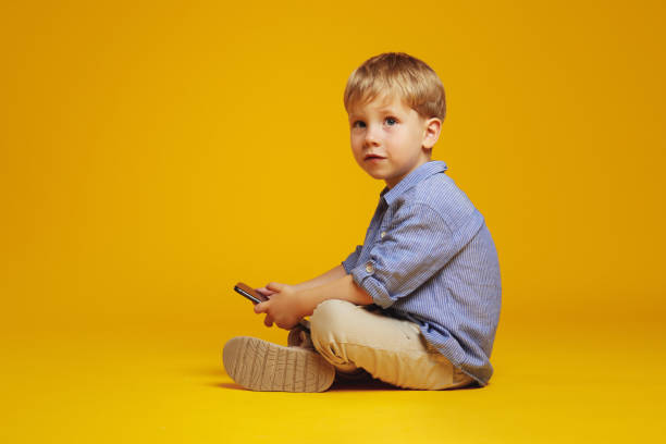istock Adorable boy sitting with crossed legs on yellow studio floor while holding mobile phone 2137967882