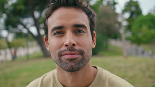 Closeup hispanic man looking camera relaxing alone in green park. Portrait handsome unshaven guy sitting on beautiful nature at weekend. Masculine carefree macho staring lens posing outdoors close up.