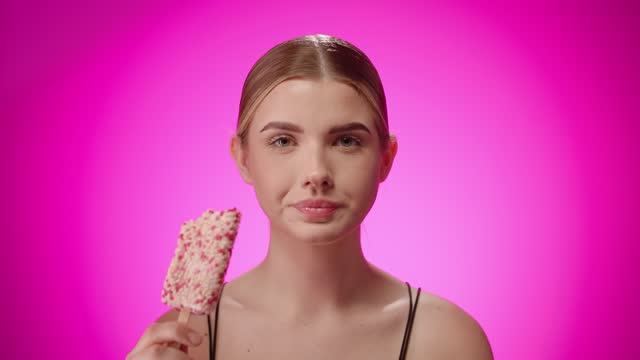 Portrait young woman bites white ice-cream popsicle, purple studio shot