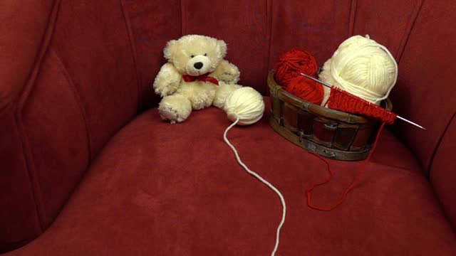 A ball of knitting thread rolls across a red velour armchair. A basket with accessories for knitting and a white teddy bear are in the armchair, slow motion