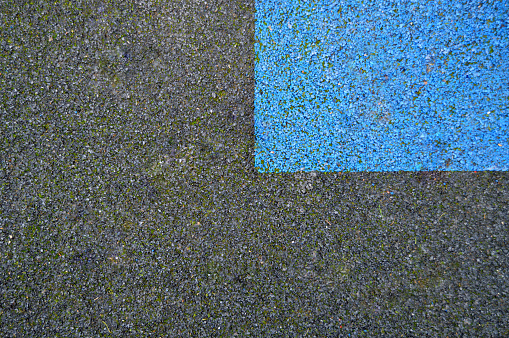 A divided granular grey black colored empty blank concrete gravel asphalt coal tar road or textured footpath background or play area pitch with a quarter or one fourth rectangular blue painted patch and green moss leaving copy space