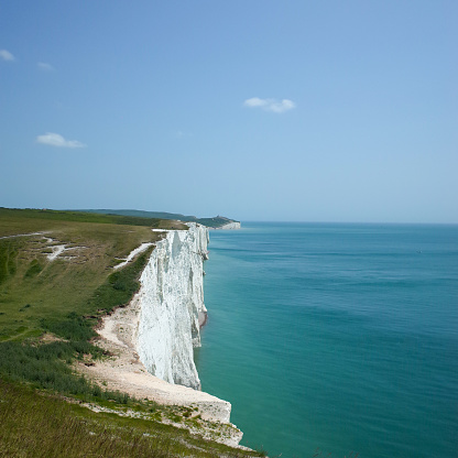A section of the white cliff taken from the top. The cliff is covered with green grass on the top. On the right is the turquoise sea. Shoot under a bright blue sky.