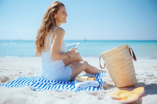 happy stylish female on the beach with straw bag and striped towel using applications on tablet PC.
