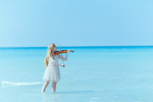 modern girl in white dress on the ocean coast with violin enjoying playing.