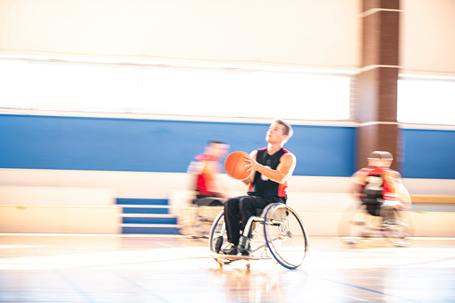 Blurred motion view of male wheelchair basketball player in mid 30s speeding in direction of basket with ball and preparing to shoot.