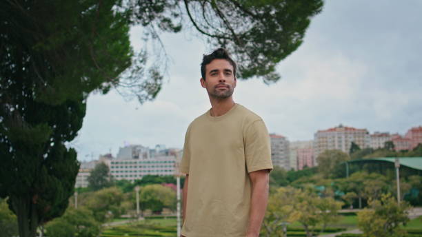 Latin guy posing green park in front cityscape close up. Handsome unshaven man looking camera standing under tree greenery alone. Smiling carefree tourist relaxing at urban weekend in town garden.