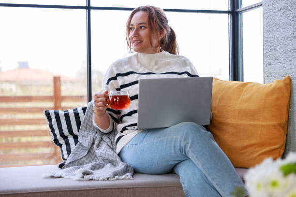 istock Young woman using a laptop computer while relaxing on the windowsill sofa at home 2138131486