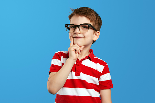 Smart thoughtful preschool boy in nerdy glasses and red striped polo shirt touching cheek and looking at camera while thinking against blue background.