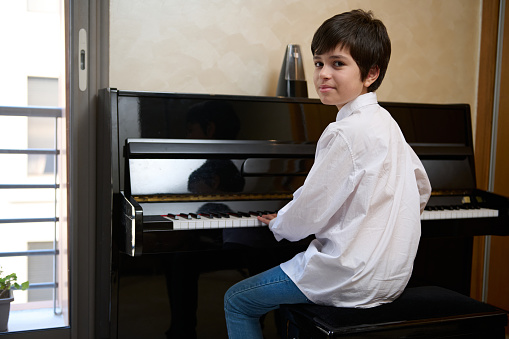 Confident teenage boy plays piano forte at home. Authentic portrait of a happy teenager student musician in white casual shirt, sitting by piano and smiling at camera, performing melody on piano forte