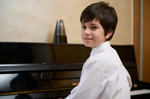 Close-up portrait multi ethnic teen boy, little pianist musician smiling looking at camera, sitting at piano forte. Handsome teenager student boy learning playing piano, during a music lesson at home