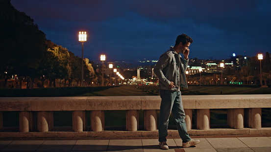 Positive guy speaking mobile phone at night city view. Smiling latin man talking smartphone at weekend urban walk. Happy carefree hipster arranging meeting by smartphone at evening town. Rest concept.