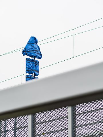 A railway sign is wrapped in blue plastic, standing erect behind the safety fence of an overpass, giving an impression of maintenance or installation work underway. The overcast sky suggests an outdoor setting during the day.