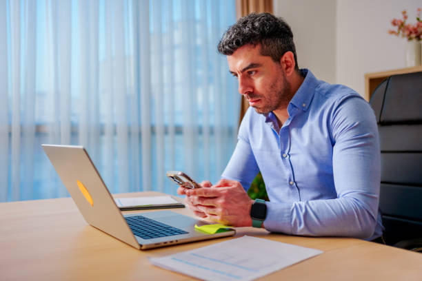 istock Confident young  man looking at smartphone while working on laptop computer in home office. Remote working, freelance, small business concept 2138250135