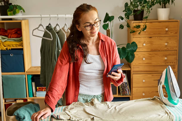 istock Happy middle aged woman using smartphone while ironing washed clothes on ironing board at home. Housewife daily routine. Housekeeping concept. 2138259118