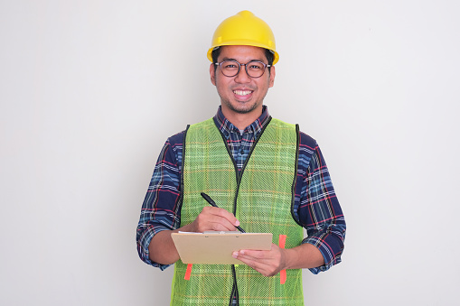 A man wearing hardhat and vest smiling while writing something on clipboard