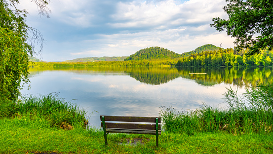 A tranquil scene of a wooden bench overlooking a calm lake, surrounded by lush greenery and hills under a partly cloudy sky. The reflection of the trees and sky in the still water adds to the peaceful ambiance.