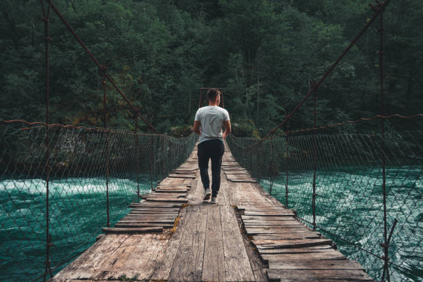 The man crossing Pedestrian Bridge over the Piva River when the dam from Piva Lake was released, creating crystal clear blue watercolor slap, surrounded by green trees on mountains
